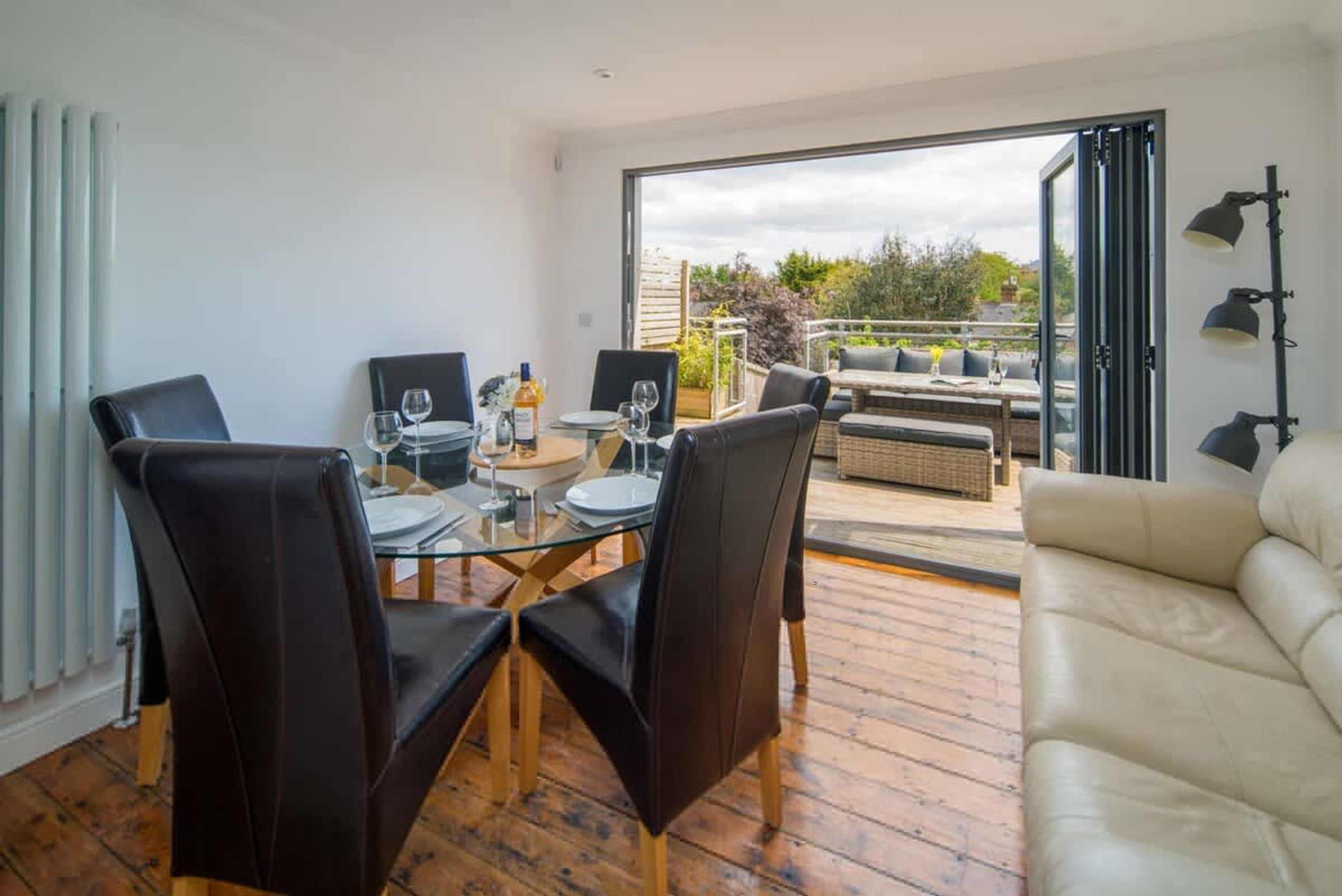 A dining area with a glass table and chairs, a sofa, and a view through a glass door to an outdoor seating area.