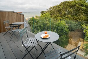 Eine Terrasse mit Tisch und Stühlen, ein Teller mit Essen und Getränken sowie Blick auf Meer und Bäume.