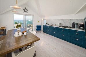 Uma cozinha moderna com uma mesa de jantar de madeira, armários azuis e vista para o mar através de grandes janelas.