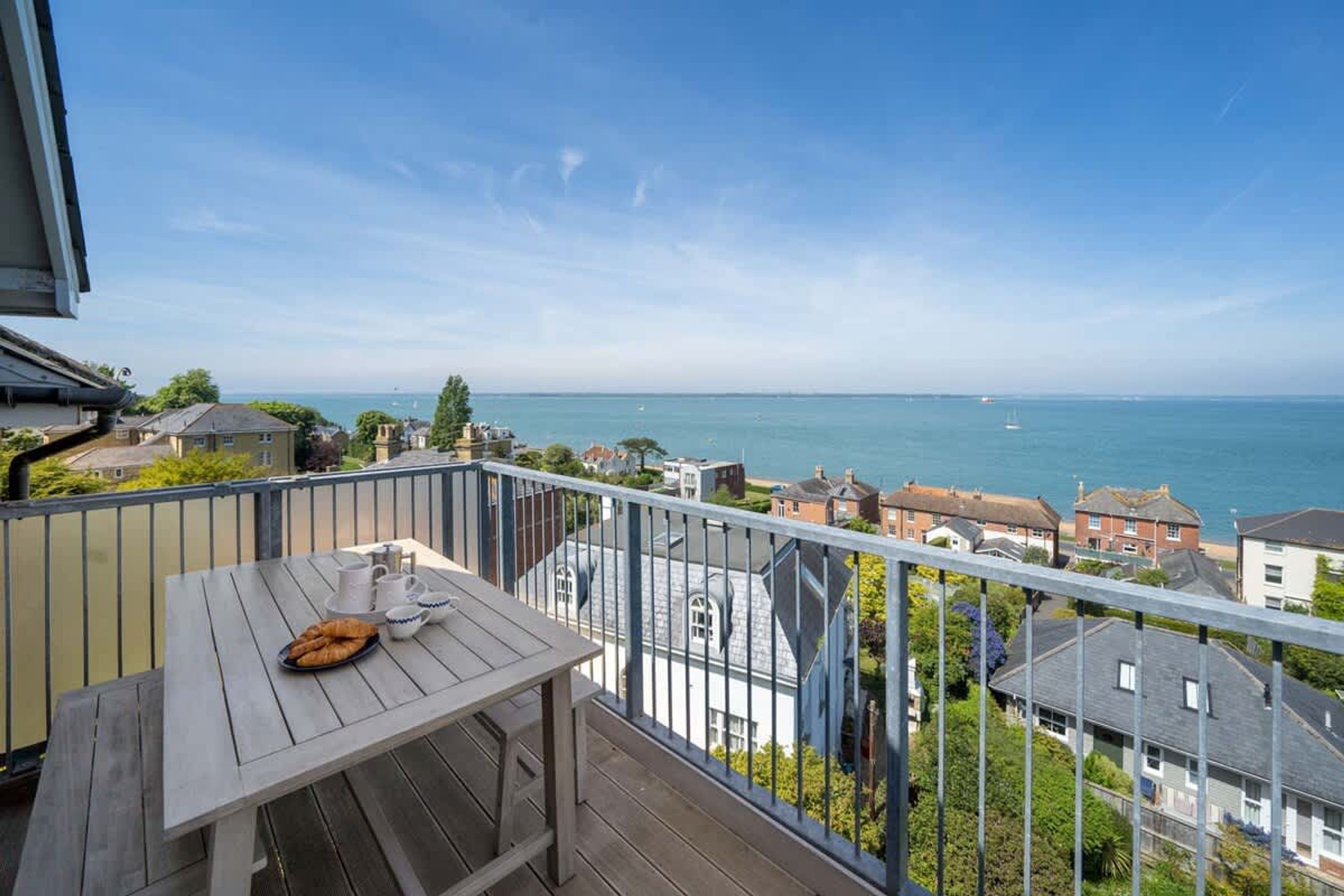 A balcony with a table, a view of the sea, and houses in the background.