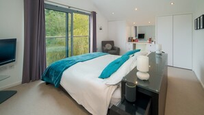 A modern bedroom with a large bed, a TV, a desk with a lamp, and a view of greenery outside. - Sandpiper House Coastal Retreat Moments From Beach (Cowes)