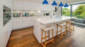 A modern kitchen with white cabinets, a central island with stools, blue pendant lights, and a view of greenery outside. - Sandpiper House Coastal Retreat Moments From Beach (Cowes)