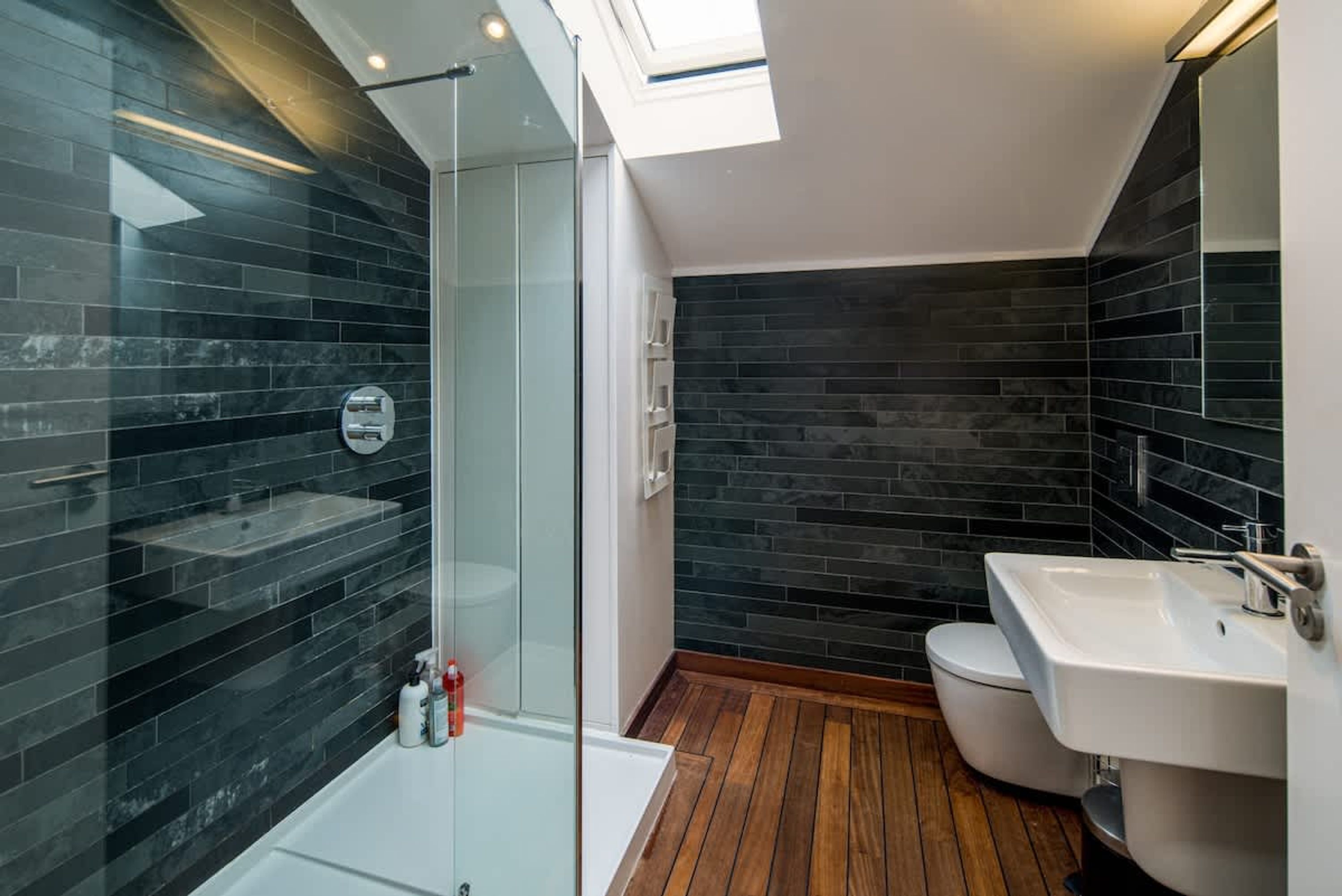 A modern bathroom with dark tiled walls, a glass shower enclosure, a white sink, and a wooden floor.