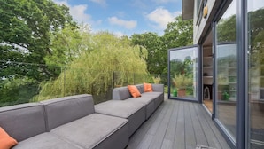 A modern outdoor deck with grey cushioned seating and orange cushions, surrounded by lush greenery. - Sandpiper House Coastal Retreat Moments From Beach (Cowes)