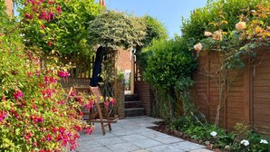 Ein Garten mit Terrassenplatten, einer Ziegelwand, einer Holzbank und verschiedenen Pflanzen.