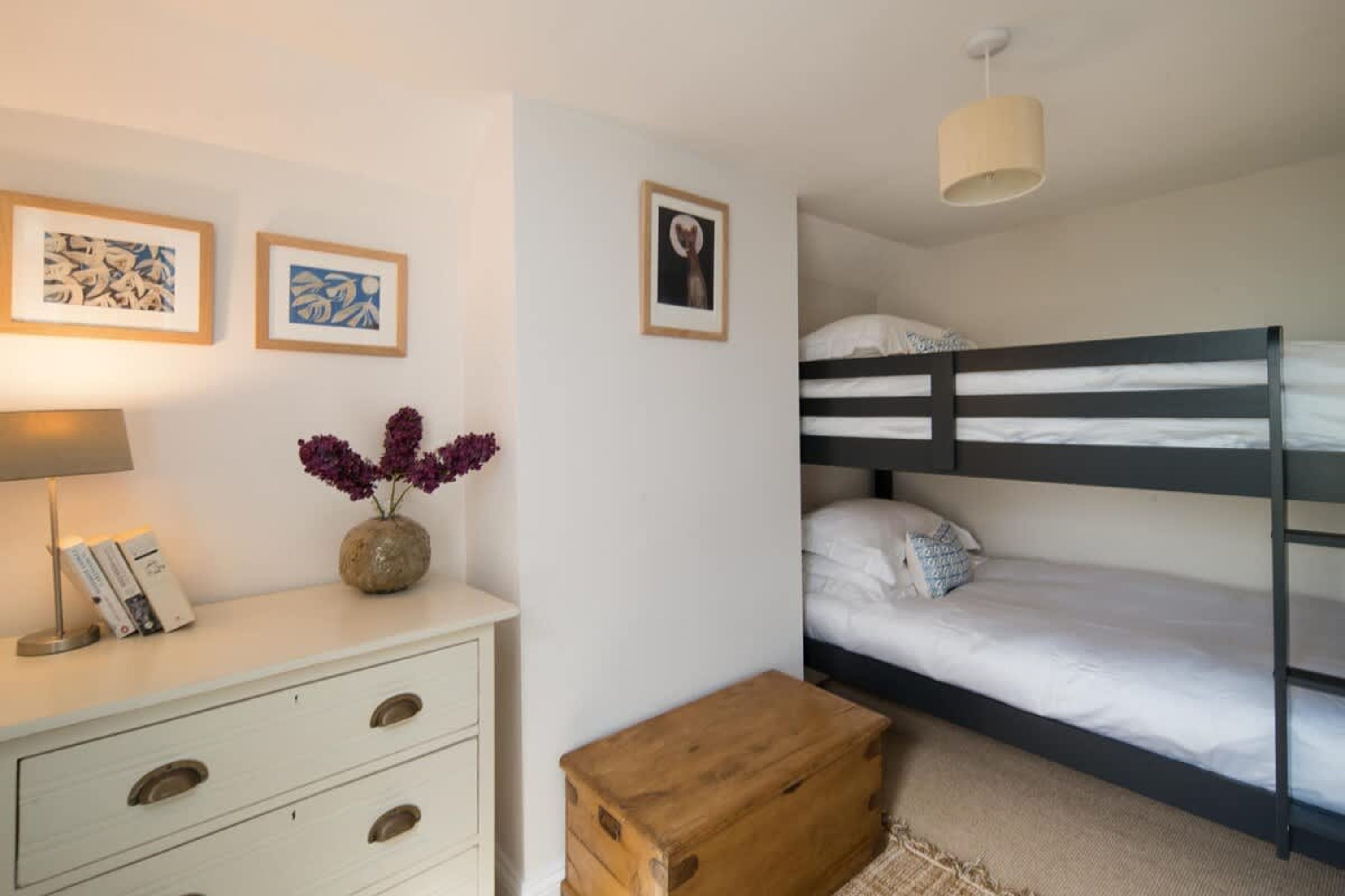A bunk bed with white bedding, a bedside lamp, framed artwork on the wall, and a small wooden chest.