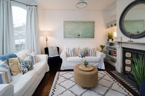 Ein Wohnzimmer mit zwei weißen Sofas, einem runden Couchtisch, einem Kamin und einem großen Fenster mit Vorhängen.