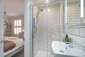 Ein Badezimmer mit Duschkabine aus Glas, weißen Fliesen an den Wänden, einem Waschbecken mit Spiegel und einem Bett im angrenzenden Zimmer.