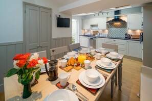 A modern kitchen with white cabinets, a flat-screen TV, and a breakfast setup with plates, cups, and a fruit basket. - Crossjack Cottage Central Cowes (Cowes)