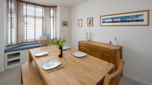 Een eethoek met een houten tafel gedekt voor vier personen, een bank met kussen en uitzicht op zee door grote ramen.
