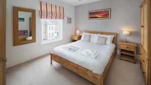 Een slaapkamer met een houten bed, een raam met gordijnen, een nachtkastje met een lamp, een spiegel en een schilderij aan de muur.