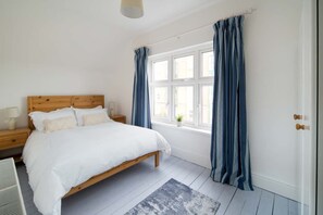A bedroom with a wooden bed, white bedding, a nightstand with a lamp, a window with curtains, and a small plant on the windowsill. - The Chambers Apartment Central Cowes (Cowes)