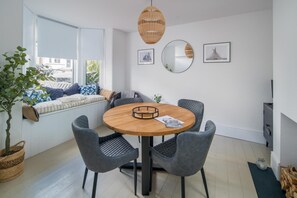 A modern living room with a round wooden table, grey upholstered chairs, a large window with a view, a hanging woven light fixture, and a small potted plant. - Wistaria Cottage Beautifully Renovated Cottage (Cowes)