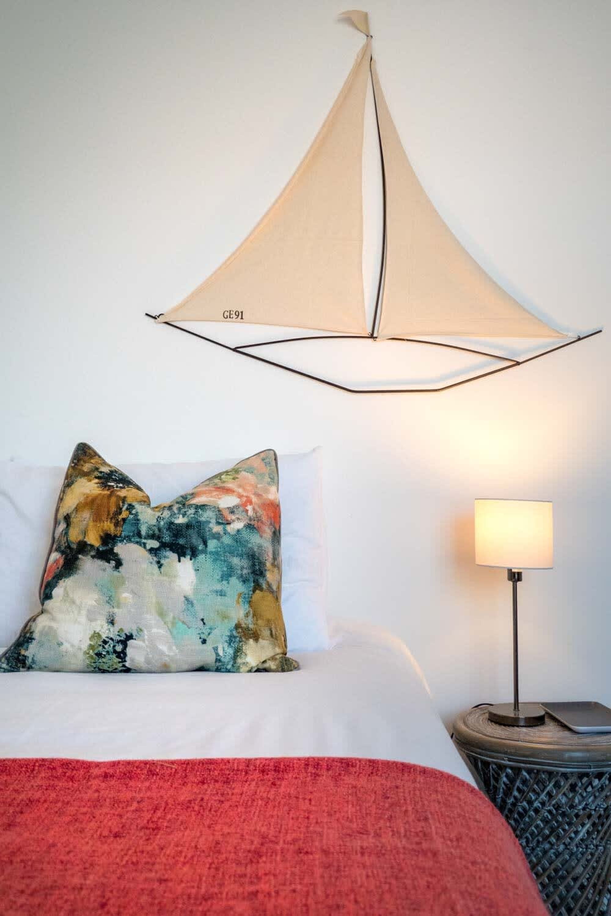 A bed with a red blanket, a pillow with a colorful abstract design, a wall-mounted canopy, and a bedside lamp.