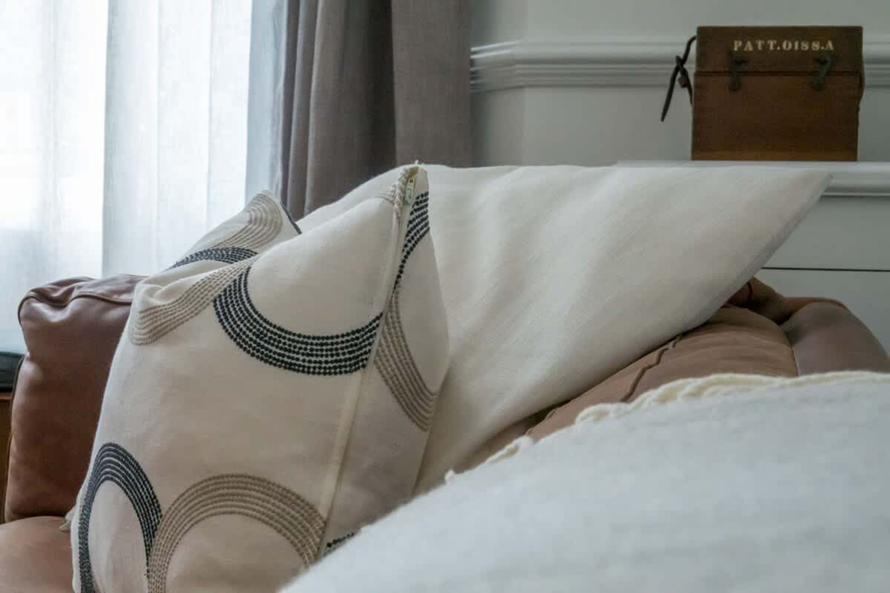 Una cama con un cojín estampado, una silla de cuero marrón y una mesita de noche blanca con una pequeña caja de madera etiquetada 'PATT.0158.A'.