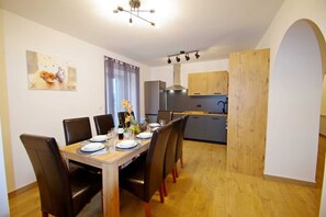 Ein Essbereich mit einem Holztisch, gedeckt für vier Personen, umgeben von Stühlen. Im Hintergrund ist eine Küche mit grauen Schränken und einer Edelstahl-Dunstabzugshaube erkennbar.