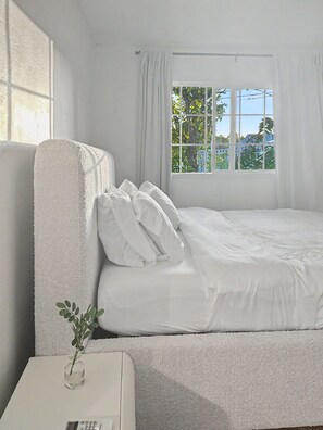 Une chambre avec un lit, une table de chevet blanche et une fenêtre avec des rideaux.