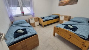 Una habitación con tres camas de madera, una ventana con cortinas ligeras, una mesita de noche de madera y un cuadro colgado en la pared, además de un piso de baldosas.
