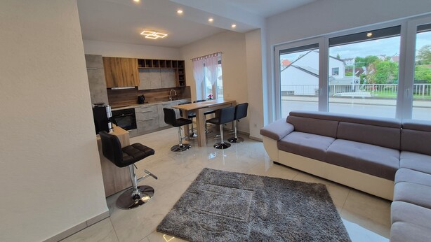 Una sala de estar moderna con un sofá, un comedor con una mesa y sillas, una cocina con armarios de madera, y un ventanal con vista a la calle.