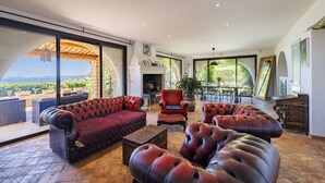 Ein großzügiger Wohnbereich mit Ledersofas, einem Kamin und großen Fenstern mit Blick ins Freie.