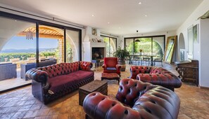 A spacious living area with leather sofas, a fireplace, and large windows offering a view of the outdoors. - Villa Le Pré Neuf – Pool & Panoramic Views (Tourrettes-sur-Loup)