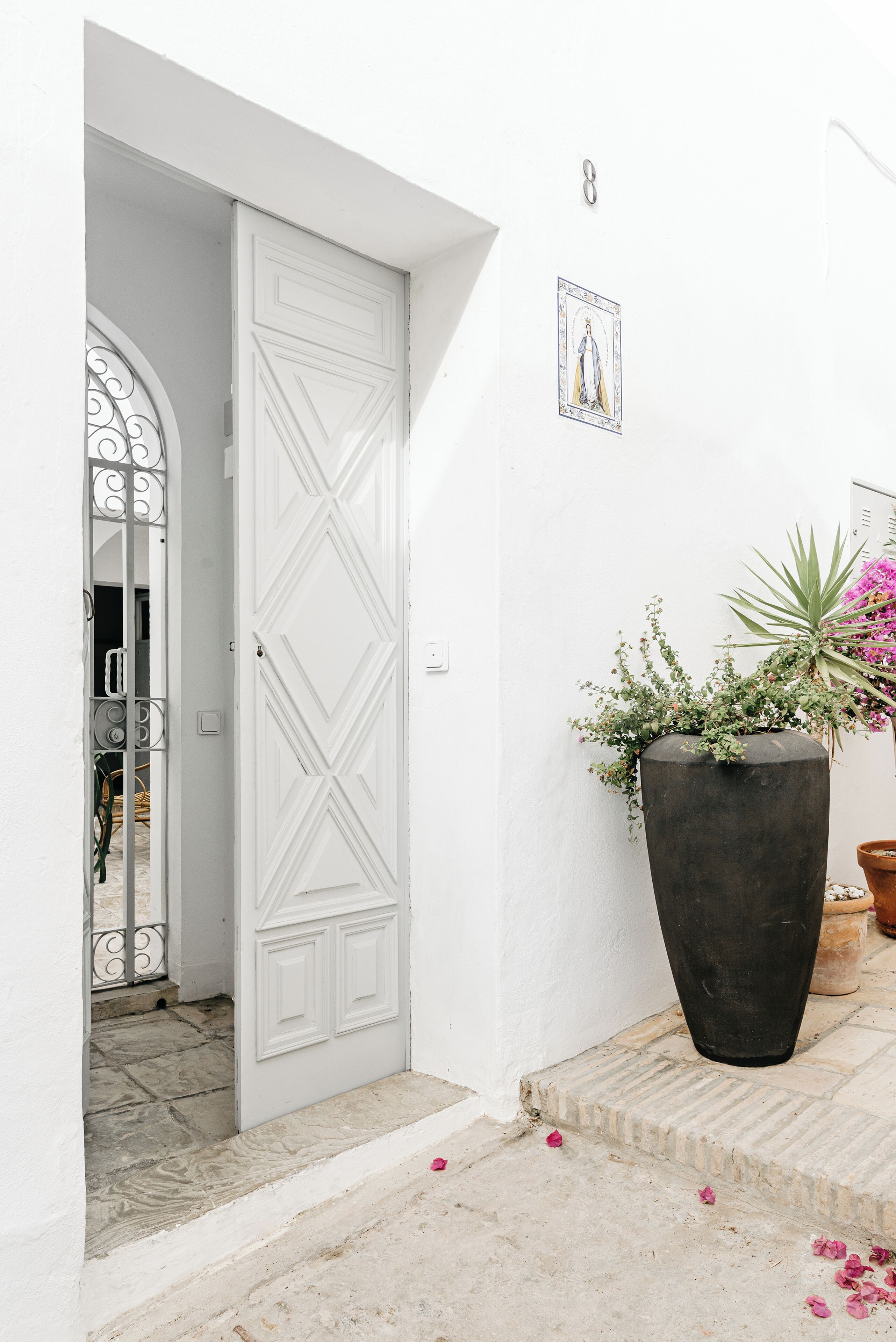 A white door with a decorative pattern, a large black planter with green plants and pink flowers, and a small framed picture on the wall.