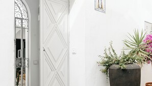 Una puerta blanca con un diseño decorativo, una maceta negra grande con plantas verdes y flores rosadas, y un cuadro pequeño en la pared.