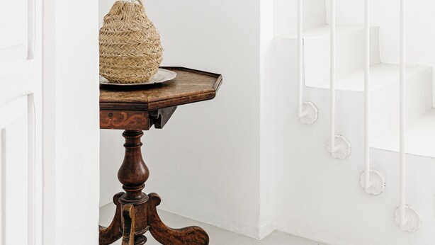 Un interior en tonos blancos con una mesa de madera y una cesta de mimbre, una escalera blanca y una barandilla también blanca.