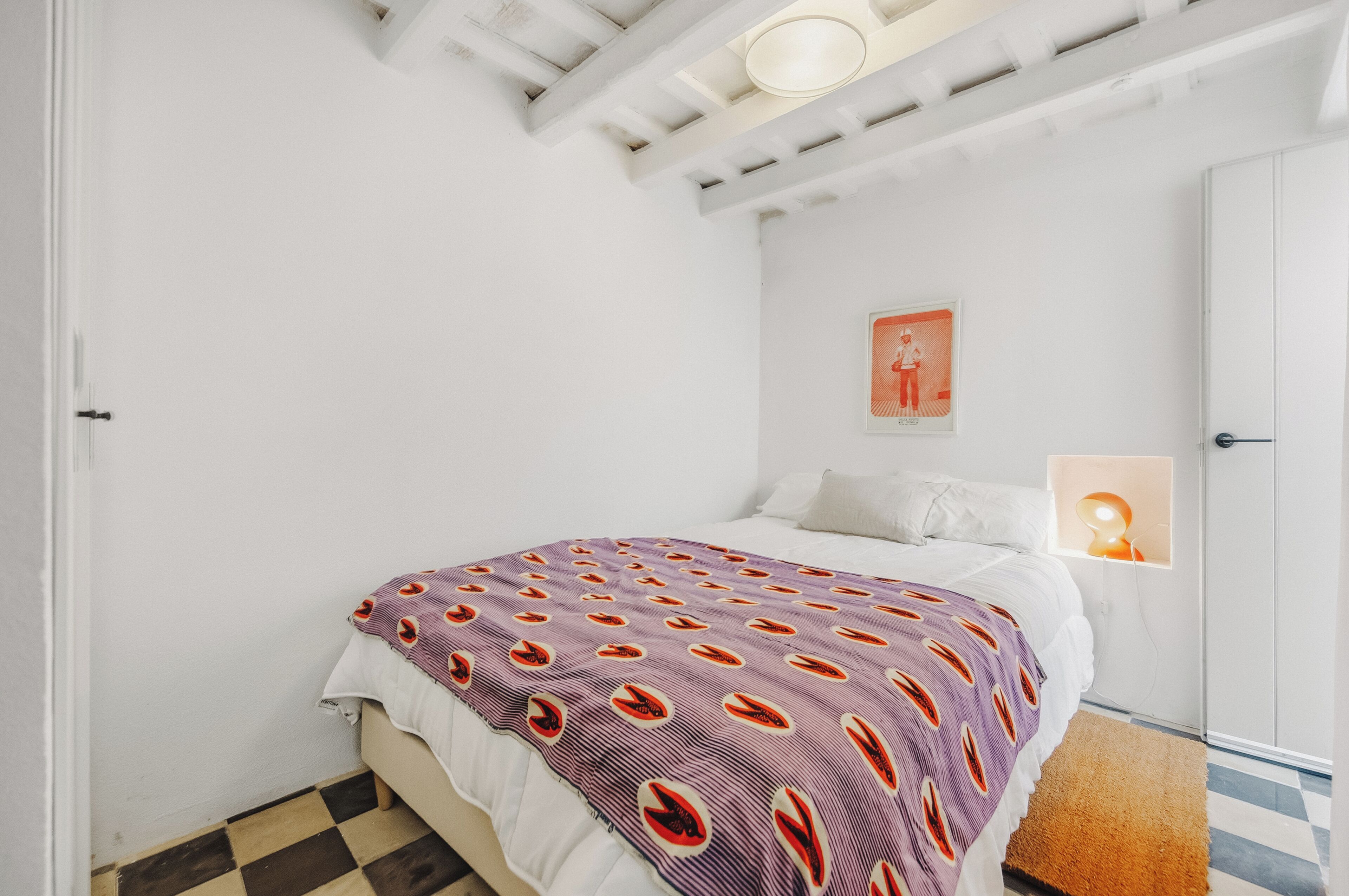 Un dormitorio con cama, una puerta blanca, un cuadro en la pared y una lámpara en el techo.