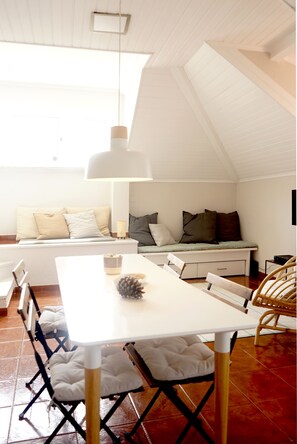 A dining area with a white table, cushioned chairs, a bench with pillows, and a ceiling light. - Old Town Apartment with Ocean Views (Sesimbra)