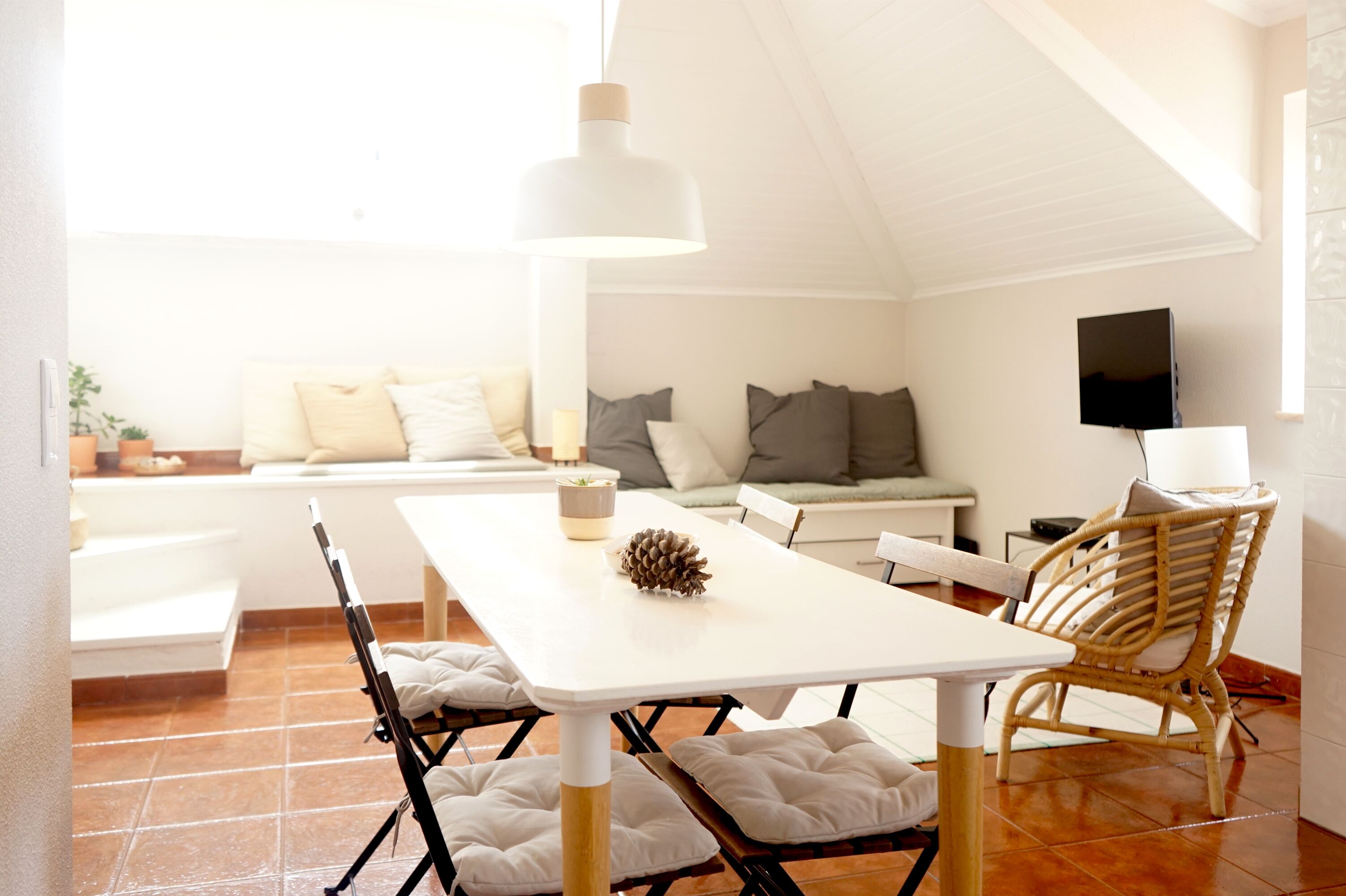 A dining area with a white table, chairs, a hanging light fixture, and a wicker chair.