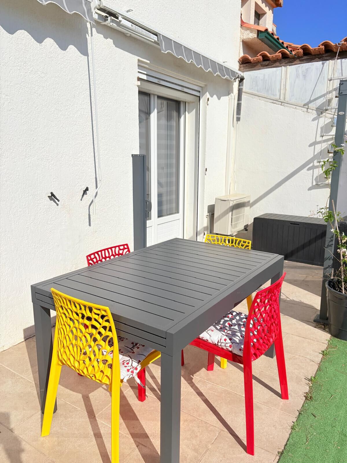 Een terras buiten met een tafel, stoelen, een deur en een kleine bloempot.