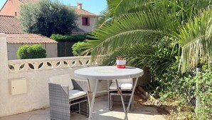Eine Terrass mit einem runden Tisch und zwei Stühlen, umgeben von Grünpflanzen und einem Gebäude im Hintergrund.
