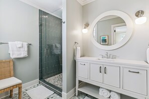 A bathroom with a shower, a white vanity with a round mirror, and a wicker chair. - Upscale East Beach & Pool (St Simons Island)