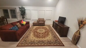Ein Wohnzimmer mit rotem Sofa, gemustertem Sessel, großem Teppich, einem Fernsehschrank und einer Topfpflanze.