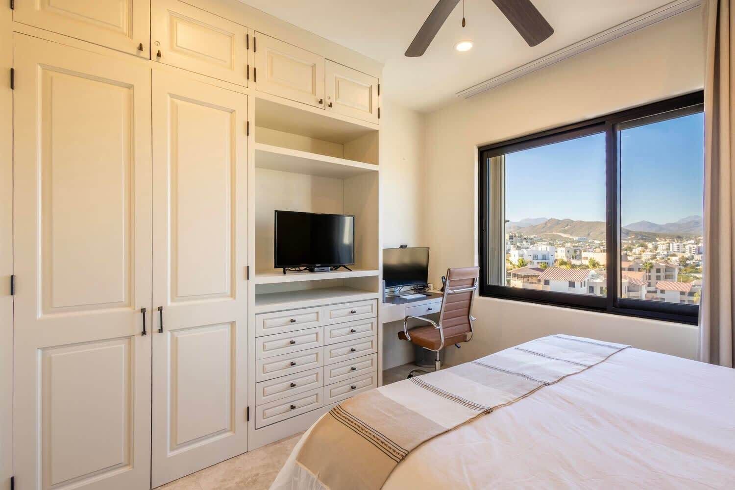 A bedroom with a bed, a desk with a chair, a TV, and a large window with a city view.