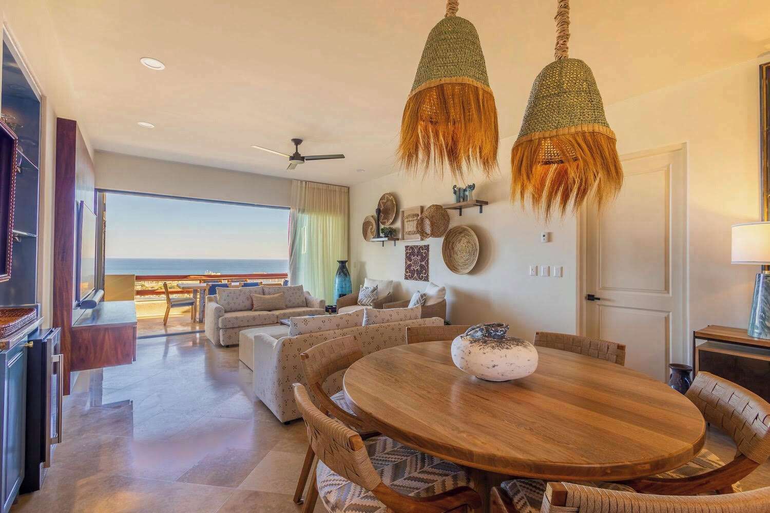 Un amplio salón con una gran mesa redonda de madera, dos lámparas colgantes y vista al océano a través de grandes ventanales.