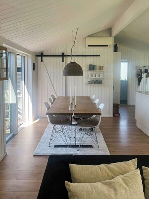Ein Essbereich mit Holztisch, weißen Stühlen, einer hängenden Pendelleuchte und einer Küche im Hintergrund.