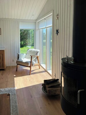 Ein Zimmer mit Holzboden, einem weißen Stuhl mit Kissen und einem großen schwarzen Heizkörper, mit Blick auf grüne Vegetation.