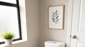 Eine weiße Toilette in einem Badezimmer mit Fenster, einer gerahmten Pflanze und einer Tür.