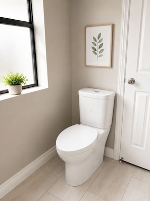 Eine weiße Toilette in einem Badezimmer mit Fenster, einer gerahmten Pflanze und einer Tür.