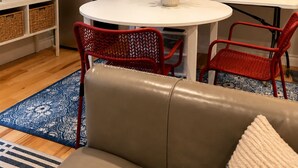 Ein kompakter Wohnraum mit einem runden Esstisch, zwei roten Stühlen, einer Couch, einem Kühlschrank und einer an der Wand befestigten Uhr.