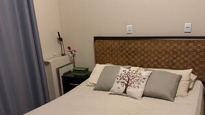Een netjes opgemaakt bed met een geweven hoofdbord, een nachtkastje met een lamp en een vaas, en een lichtschakelaar aan de muur.
