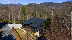 Ein modernes, zweistöckiges Haus mit dunklem Dach, umgeben von einer bewaldeten Landschaft und Bergen im Hintergrund.