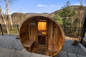 Eine Holzsauna mit Bänken im Inneren, umgeben von einem Metallzaun, vor einer Berglandschaft.