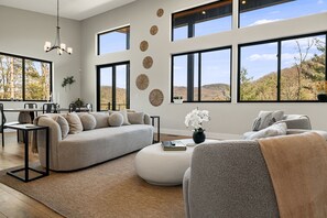 Ein modernes Wohnzimmer mit einer Couch, einem Hocker und Sesseln, großen Fenstern mit Blick auf die Hügel und einer Kristallleuchtre.