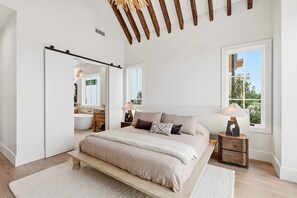 A bedroom with a large bed, a sliding door to a bathroom, a window with a view, and a wooden ceiling beam.