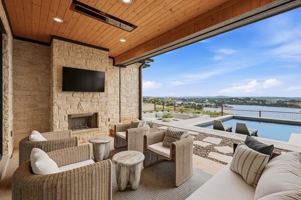 A stone fireplace, wicker furniture, a flat-screen TV, and a pool with a view of a lake and distant buildings.