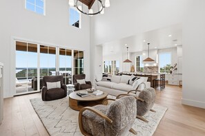 A spacious living room with a large window, a sofa, two armchairs, a coffee table, and a kitchen area in the background.