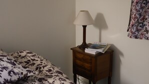 Un letto con copriletto a motivi floreali, un comodino di legno con una lampada, un libro e una parete con tenda.